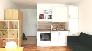 A compact kitchen with white cabinets, a wooden countertop, and a built-in oven. There is a green sofa and a wooden table with baskets in the living area. - Rafael Kaiser - Apartment Sunlight (Graz)