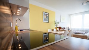 A modern kitchen with a glass-top island, a built-in oven, and a stainless steel sink. There is a dining area with a white table and chairs, and a large window providing natural light. - Rafael Kaiser - Apartment Sunlight (Graz)