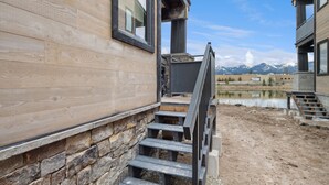 Fachada de madera con escalones de piedra que conducen a un balcón con vistas a un lago y montañas.