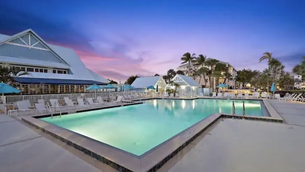 A large outdoor swimming pool with lounge chairs and umbrellas, surrounded by palm trees and buildings with white roofs.