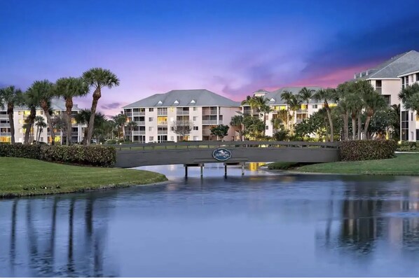 A hotel with a bridge over a calm body of water, surrounded by palm trees and a clear sky.