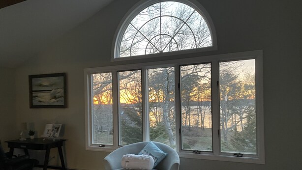 A room with a large arched window, a round ottoman, a chair, a small table with a lamp, and a framed picture on the wall. - Buzzards bay hide away (Bourne)