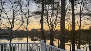A wooden deck with white railings overlooking a calm lake, surrounded by trees and a distant house. - Buzzards bay hide away (Bourne)