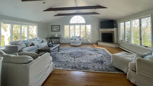 A spacious living room with a high ceiling, large windows, a fireplace, and a cozy seating area. - Buzzards bay hide away (Bourne)