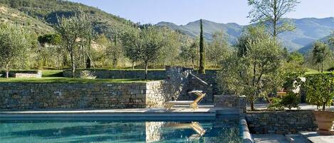 A pool area with stone walls and steps leading into the water, surrounded by trees and a mountainous backdrop.