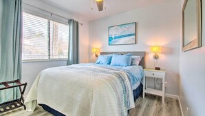 Een slaapkamer met een bed, een plafondventilator, een nachtkastje met een lamp, een raam met gordijnen en een schilderij aan de muur.