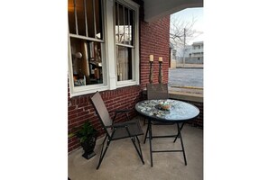Eine Terrasse mit rundem Tisch und zwei Stühlen, ein Fenster mit Blick auf die Straße und eine Ziegelwand.