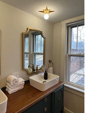 Ein Badezimmer mit einem weißen Waschbecken, einem Spiegel, einem Stapel Handtücher und einem Fenster mit Blick auf Bäume.