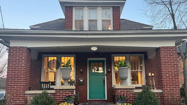Una casa de ladrillo de dos pisos con una puerta verde, dos ventanas grandes con plantas en macetas y un pequeño porche.
