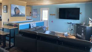 Un salon avec un plafond en bois, un grand téléviseur à écran plat, un canapé, une table basse et un coin cuisine doté de meubles en bois et d’un tableau encadré.