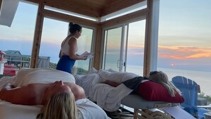 Une cabane en bois dotée de grandes fenêtres donnant sur une étendue d’eau. À l’intérieur, on peut voir deux lits où sont allongées des personnes.