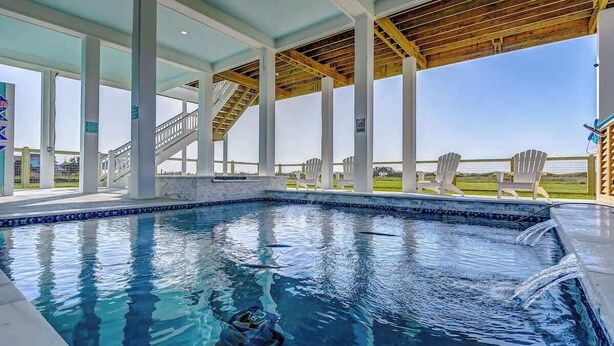 A covered swimming pool with a wooden ceiling, clear blue water, and a view of a grassy area with chairs.