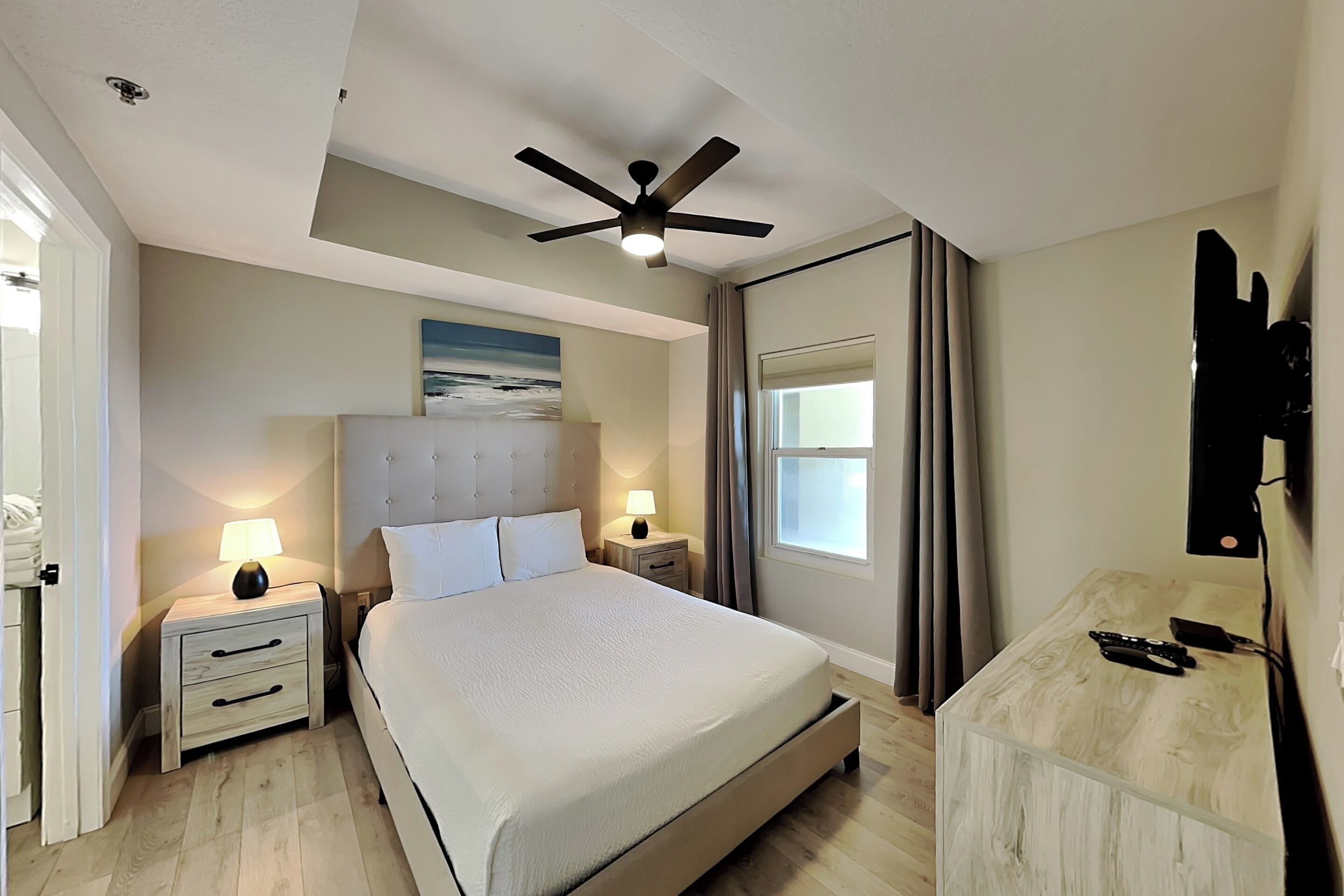 A bedroom with a bed, bedside tables, a ceiling fan, a window with curtains, and a painting on the wall.