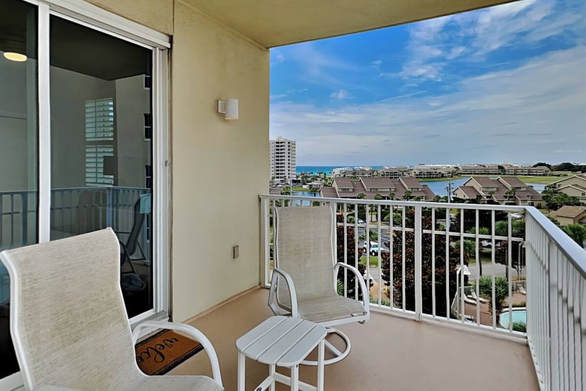 A balcony with a white chair and table, a sliding glass door, and a view of a residential area with buildings and a body of water.