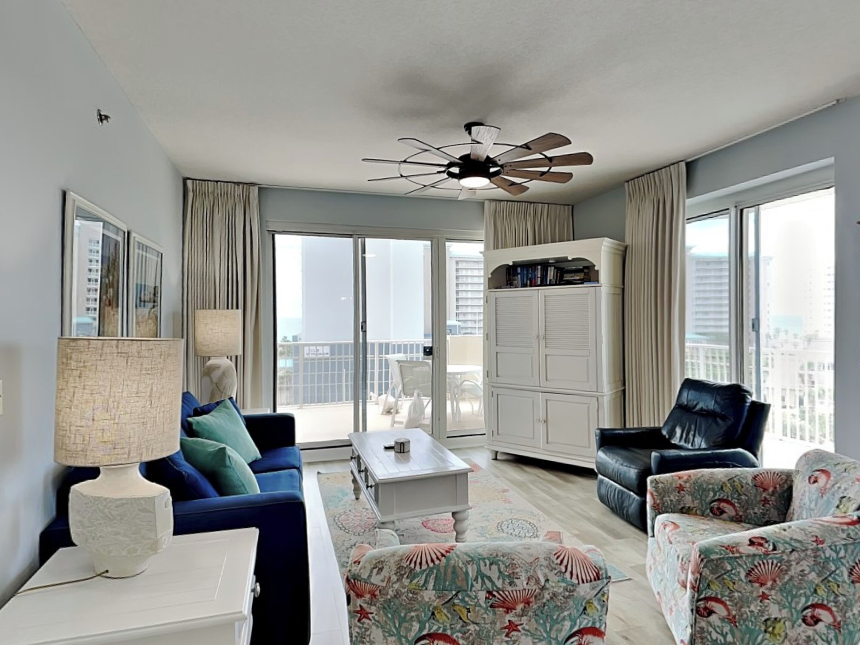 A modern living room with a blue sofa, a white coffee table, a black armchair, and a patterned ottoman. There is a ceiling fan, a large window with curtains, and a view of the city.