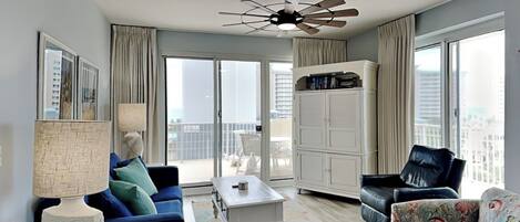 A modern living room with a blue sofa, a white coffee table, a black armchair, and a patterned ottoman. There is a ceiling fan, a large window with curtains, and a view of the city.