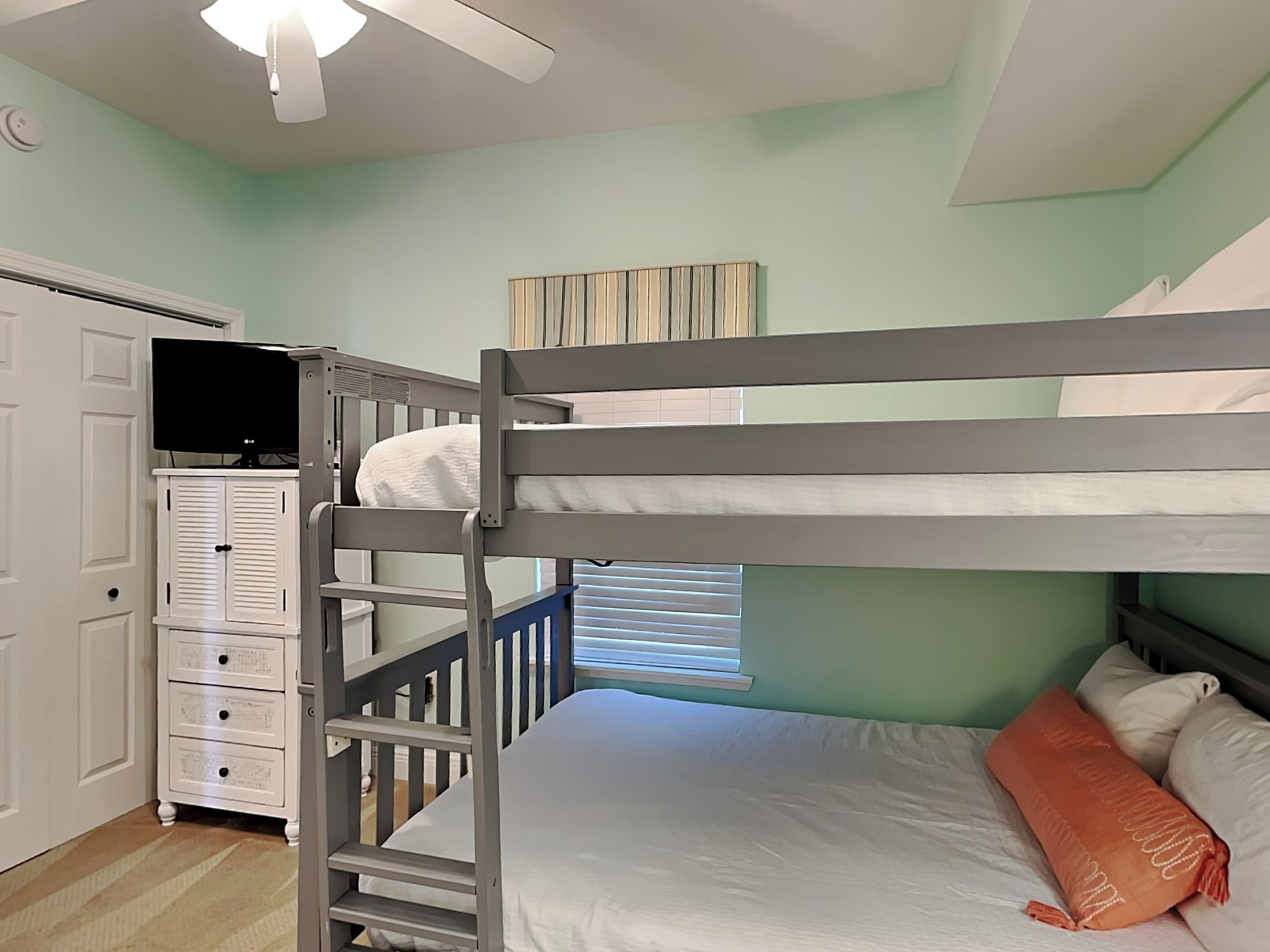 Un lit superposé avec un téléviseur placé sur le lit du haut, une armoire blanche et une fenêtre avec des stores.