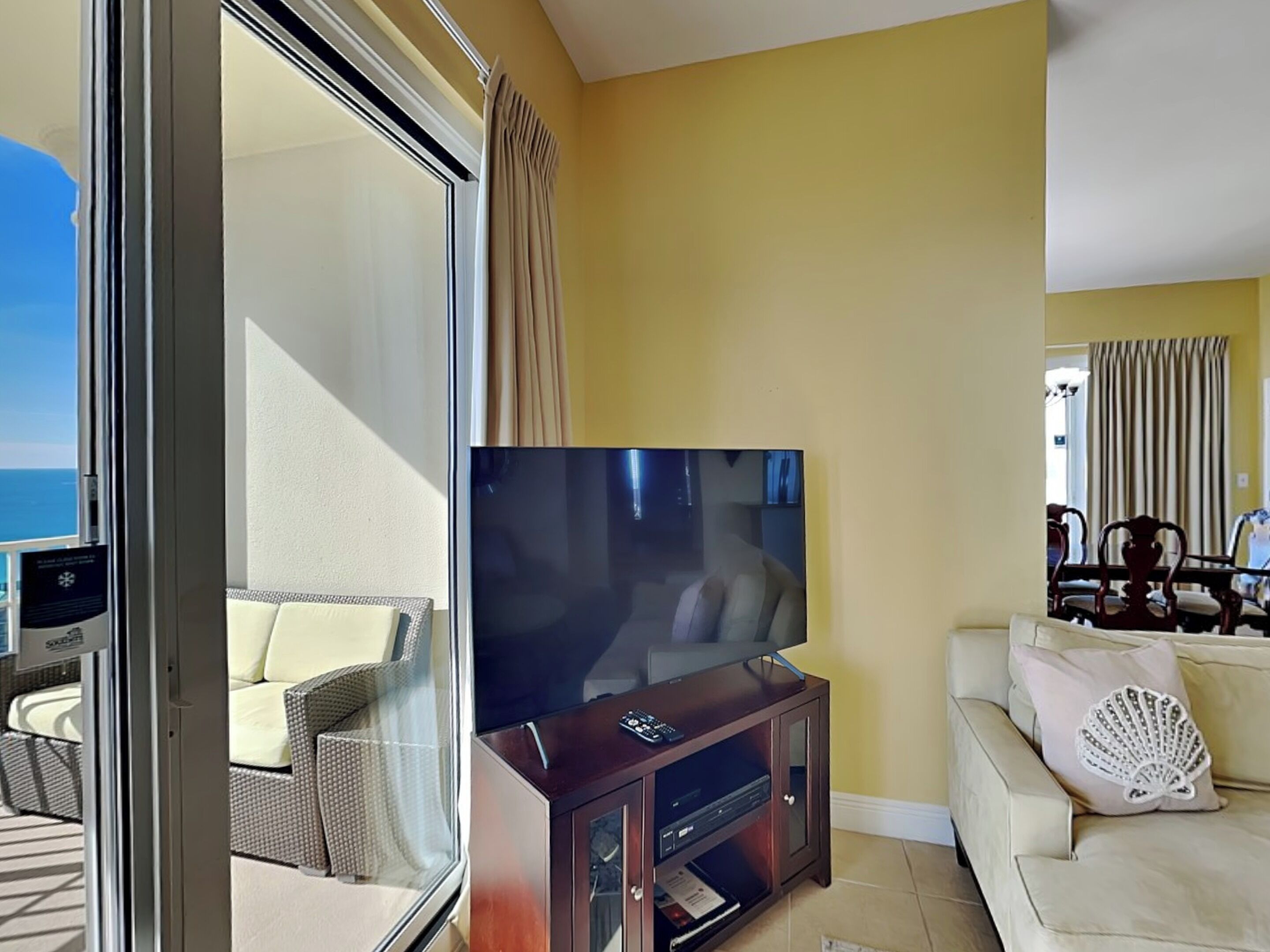 Una sala de estar con una televisión de pantalla plana grande sobre un soporte de madera, un sofá color beige y un comedor con sillas y mesa de madera.