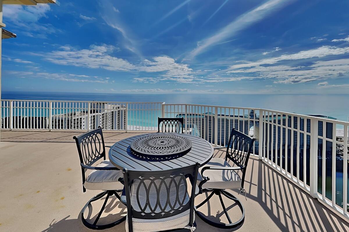 A round table with four chairs on a balcony overlooking a beach.