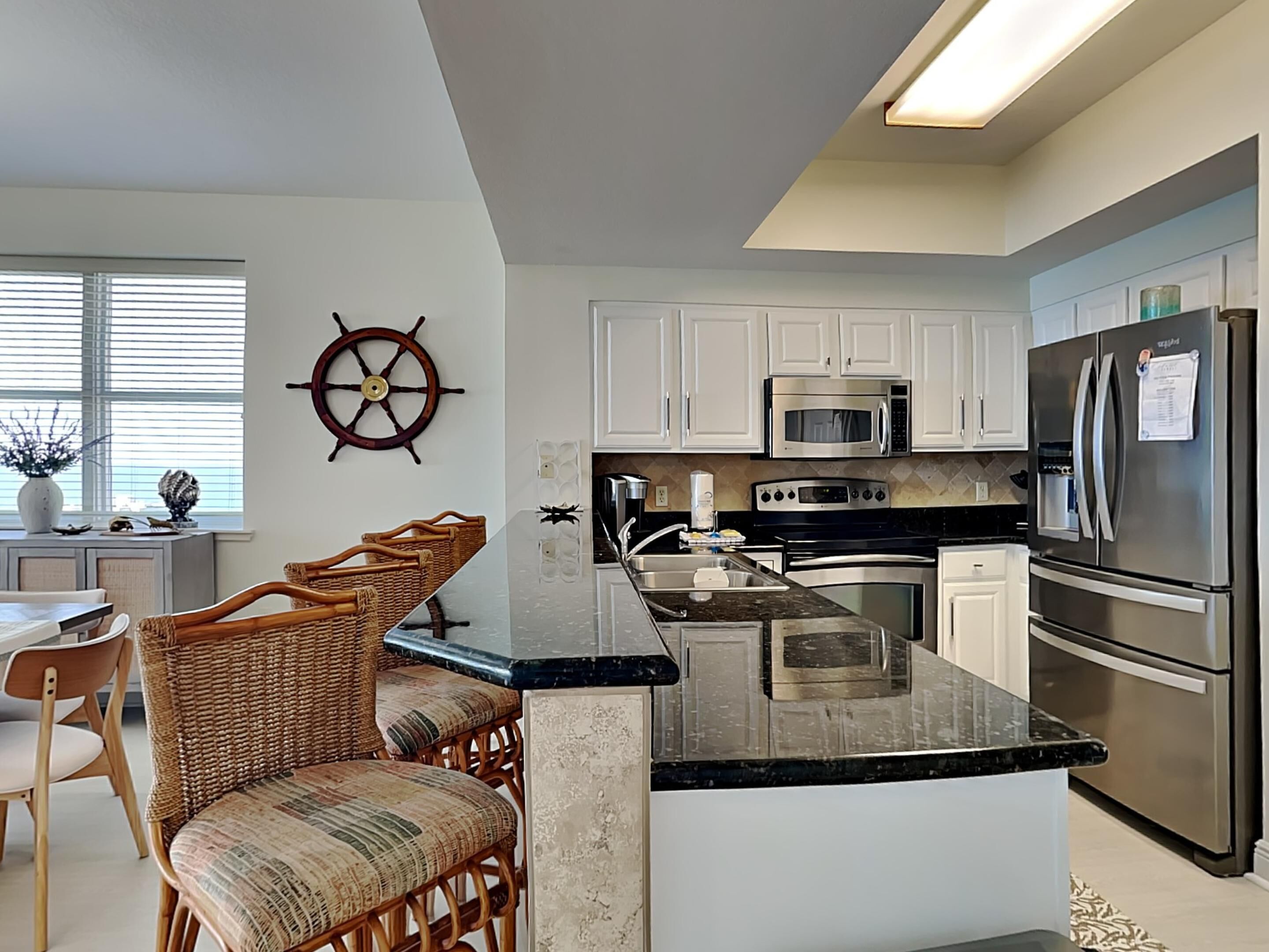 A modern kitchen with white cabinets, stainless steel appliances, a large island with a black countertop, and a ship's wheel decoration on the wall.
