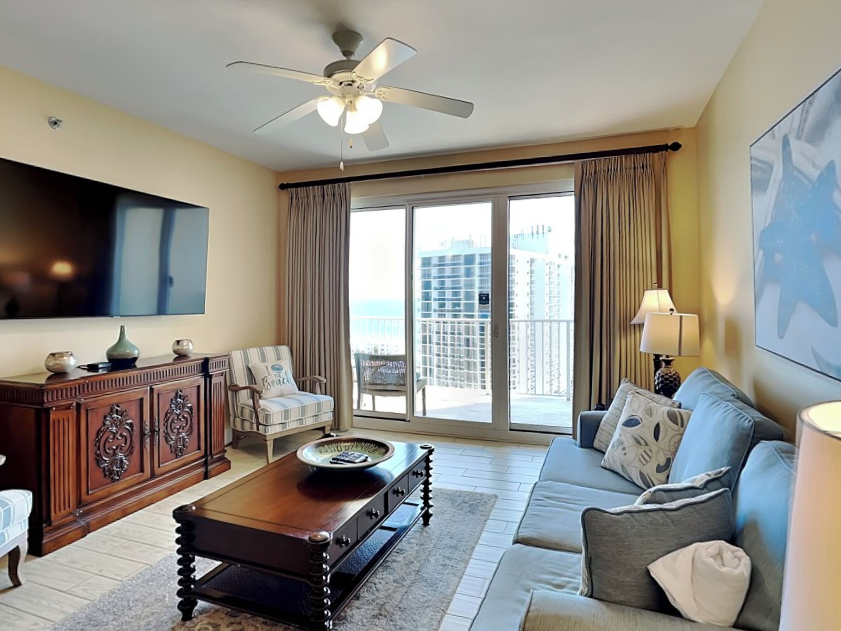 A spacious living room with a large flat-screen TV, a wooden coffee table, a sofa, and a chair. There is a balcony with a view of high-rise buildings.