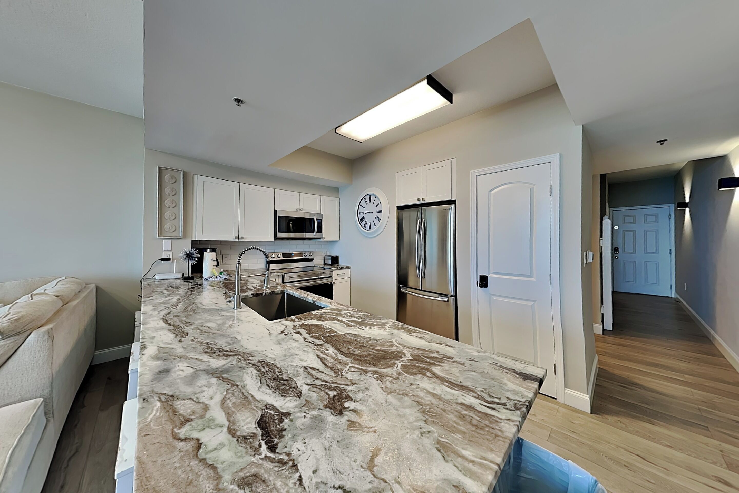 A modern kitchen with white cabinets, a marble countertop, stainless steel appliances, and a clock on the wall.