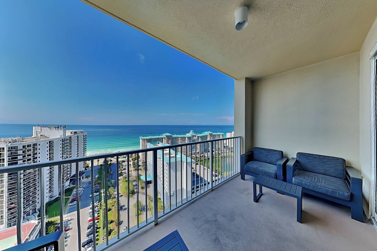 A balcony with a view of the ocean, a seating area with two sofas and a table, and a high-rise building in the background.