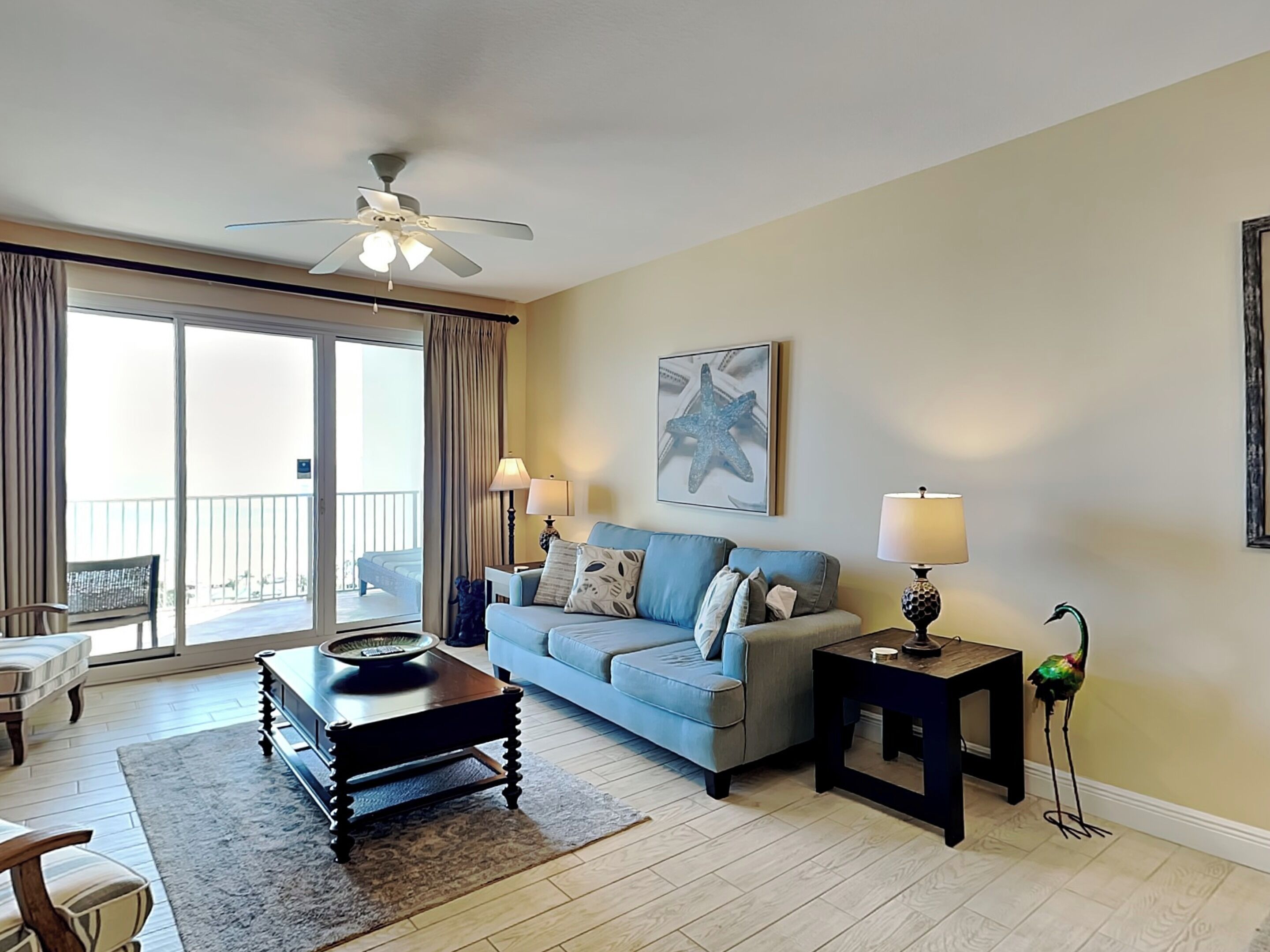 A living room with a sofa, coffee table, and lamps. There is a balcony with a bed and a chair.