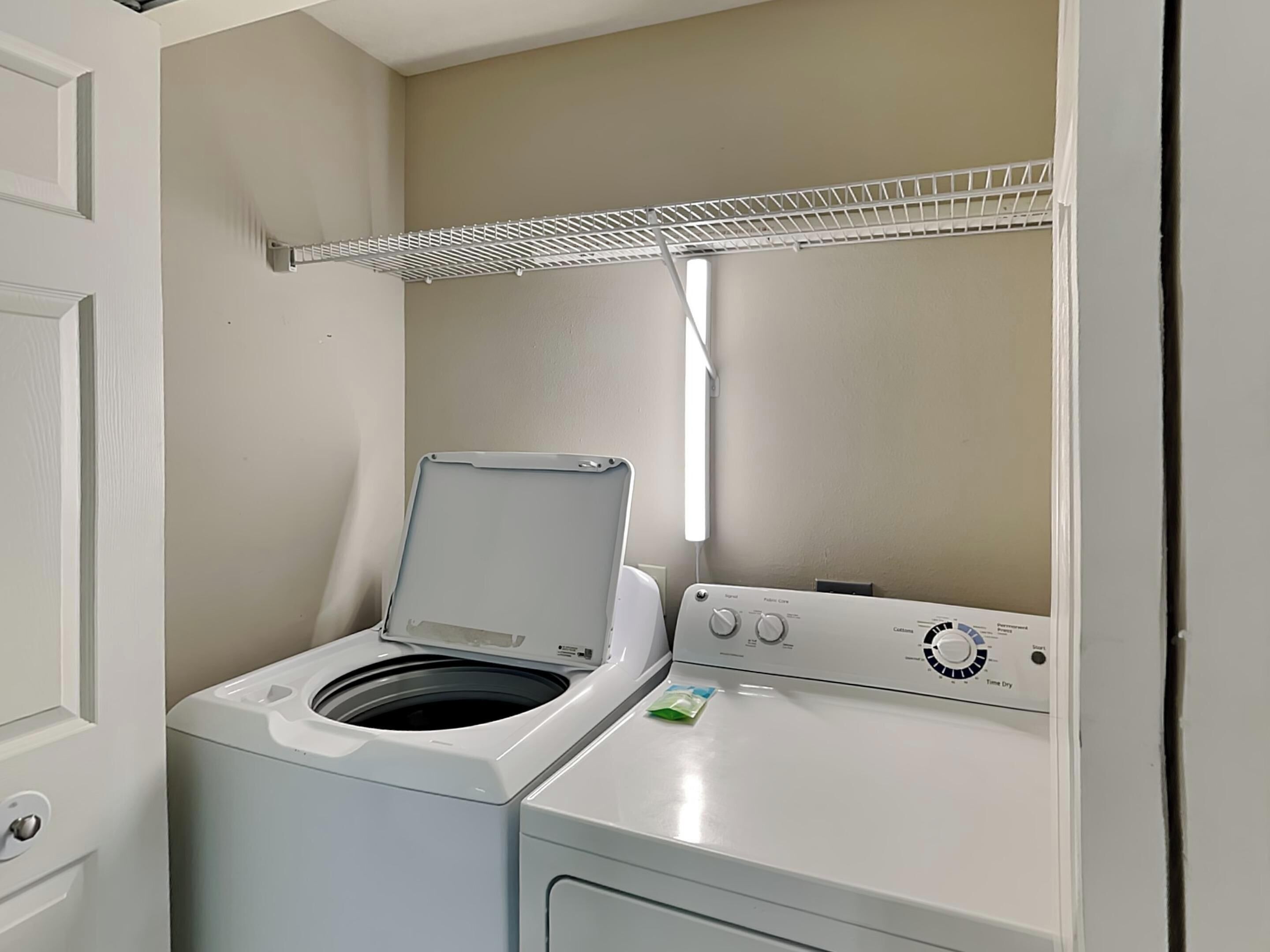 Un espace buanderie compact comprenant un lave-linge et un sèche-linge à chargement frontal, un étendoir métallique et une porte située à gauche.