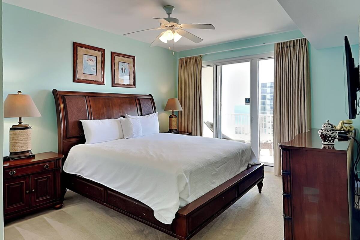 A hotel room with a large bed, two bedside tables with lamps, a ceiling fan, and a view of the city through the balcony doors.