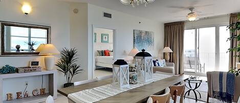 A modern living room with a dining area, featuring a long wooden table, comfortable seating, a chandelier, and a large window with a view.