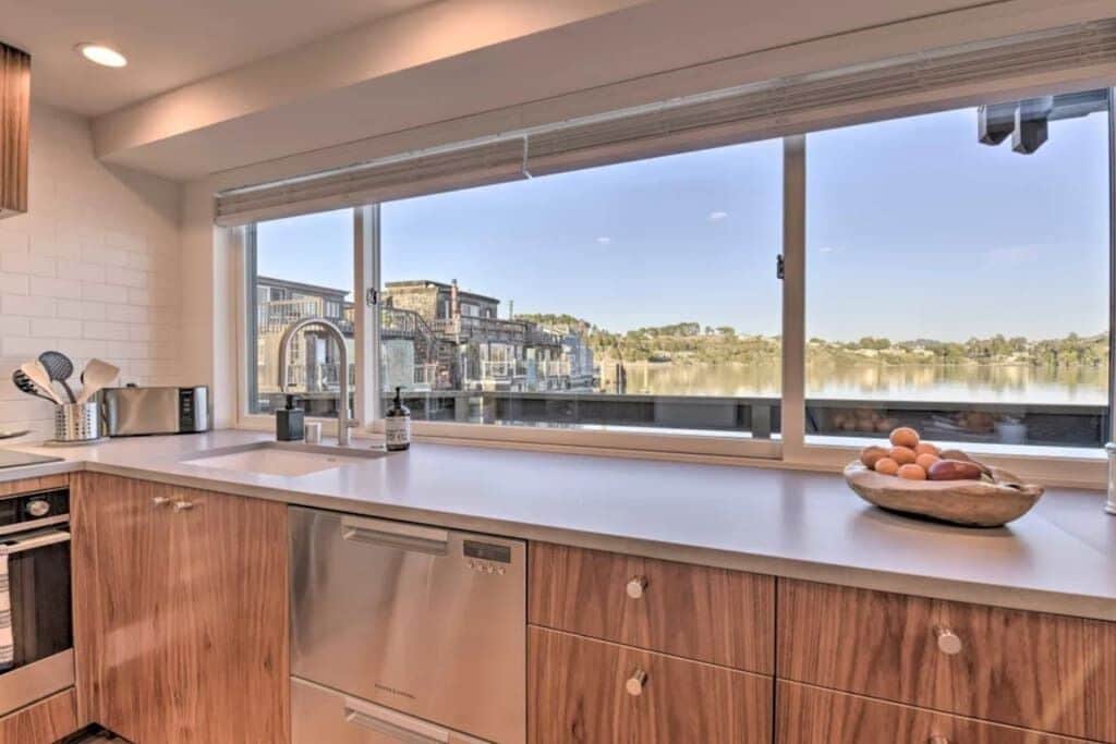 Cocina moderna con armarios de madera, electrodomésticos de acero inoxidable y un amplio ventanal con vistas a un edificio y árboles.