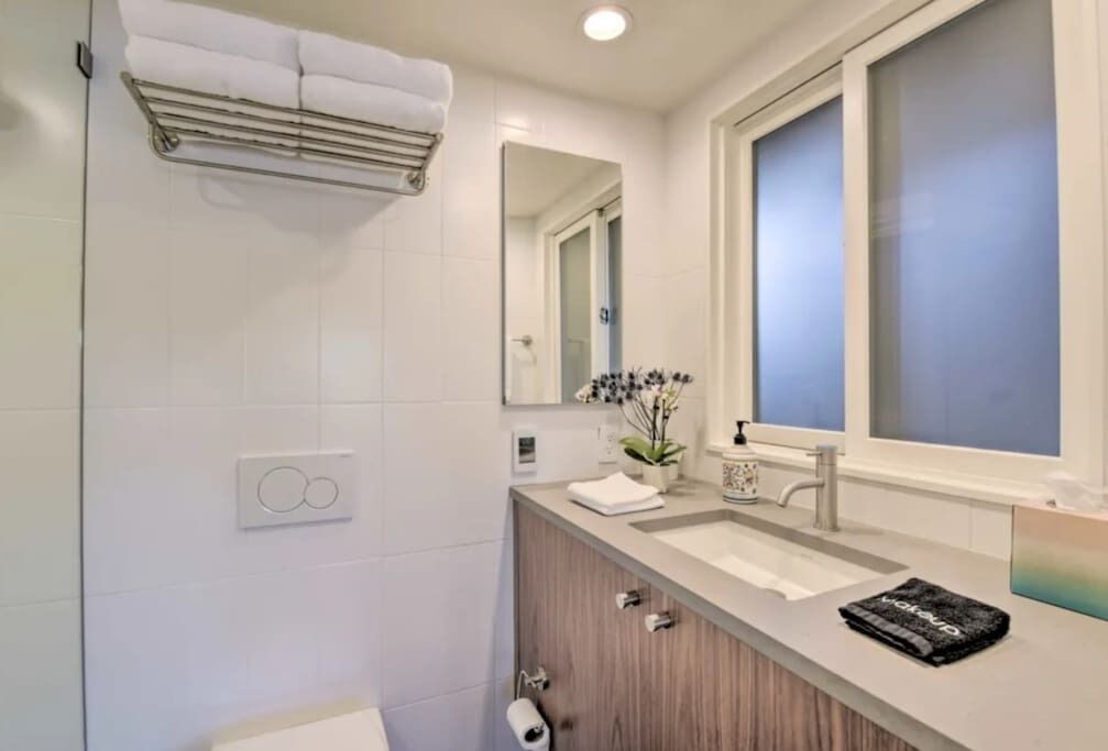 A modern bathroom with a white tiled wall, a wooden vanity, a sink, a mirror, and a towel rack.