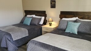 Deux lits jumeaux avec des couvertures grises et des coussins turquoise, une table de chevet avec une lampe, et un mur orné d’une citation encadrée.
