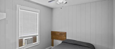A bedroom with a bed, a ceiling fan, a window with blinds, and an air conditioning unit.