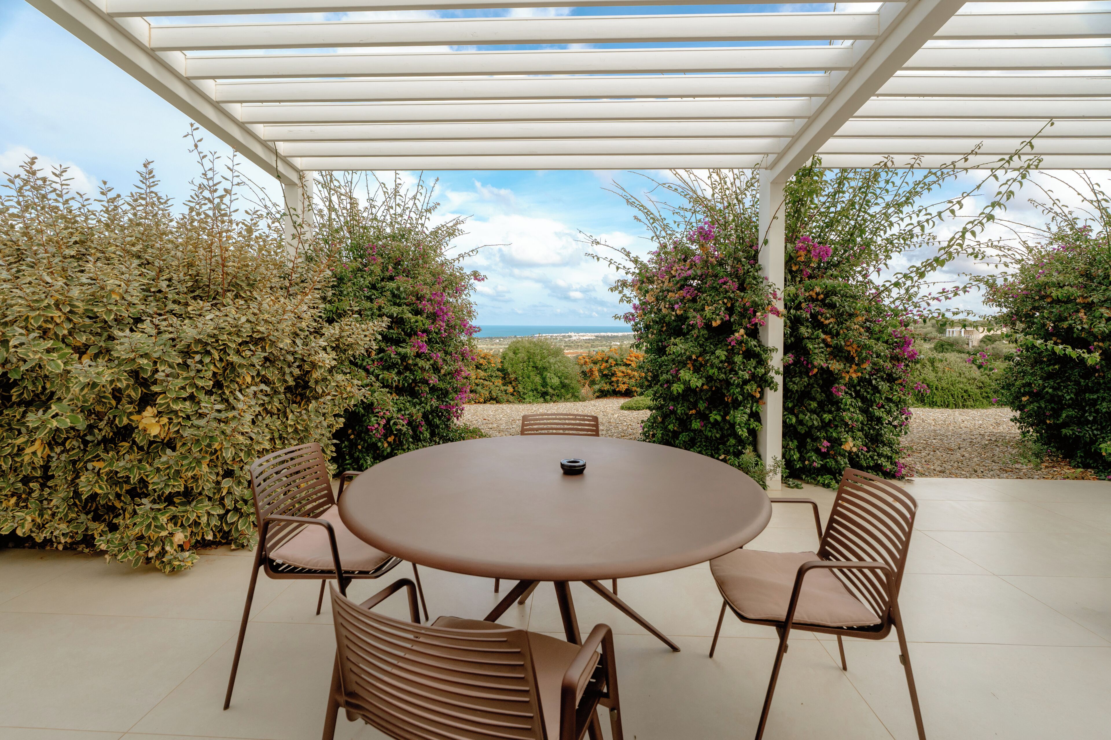 Een overdekt terras met een ronde tafel en stoelen, omringd door weelderige begroeiing en uitzicht op het landschap.