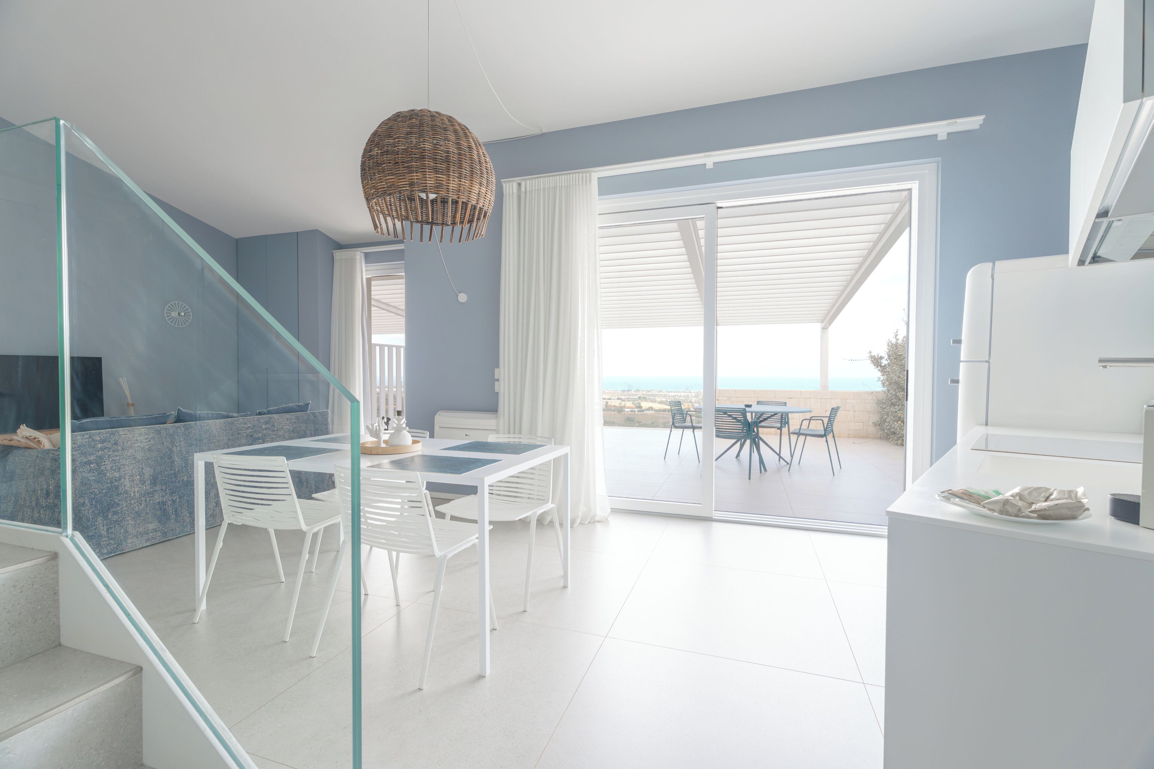 Een modern interieur met een glazen trap, een eetgedeelte met witte stoelen en een tafel, en uitzicht door een glazen deur op een terras.