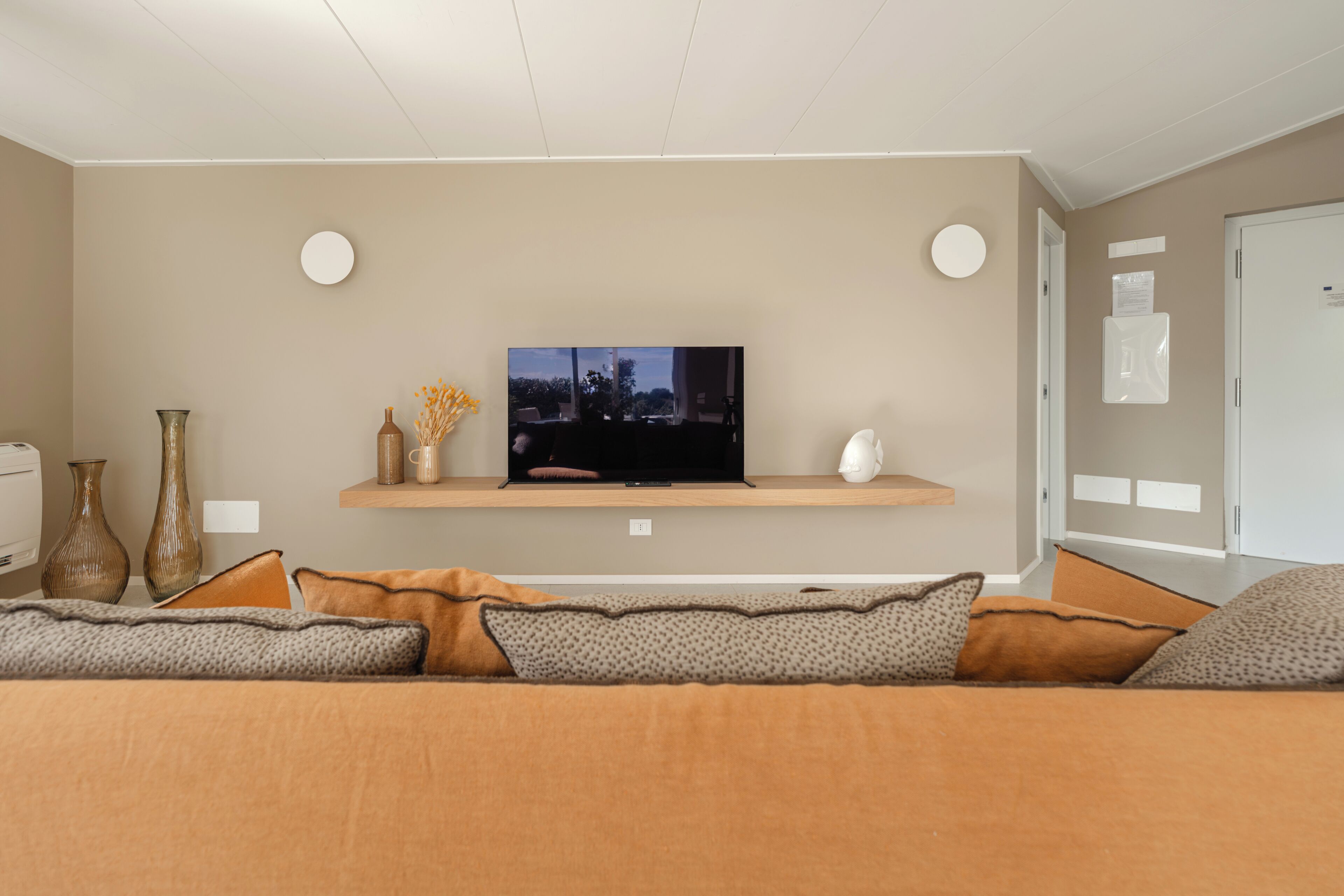 Una sala de estar moderna con un televisor de pantalla plana sobre una estantería de madera, un sofá con cojines estampados y una puerta con pomo blanco.