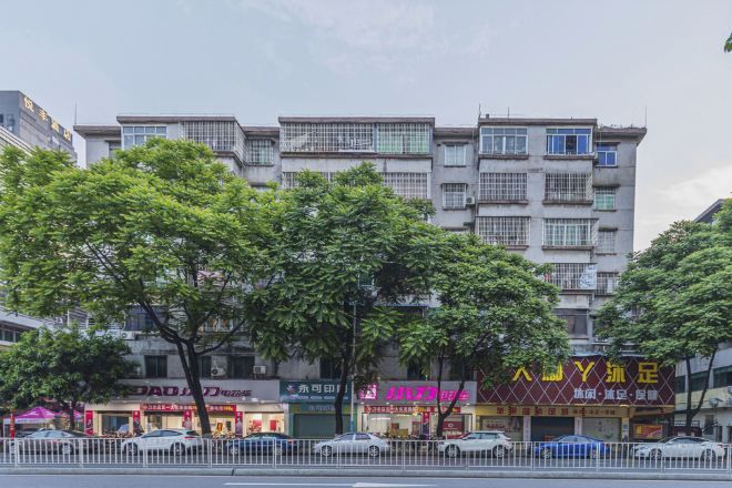 Un edificio de varios pisos con comercios en la planta baja, una cerca blanca en primer plano y árboles al fondo.