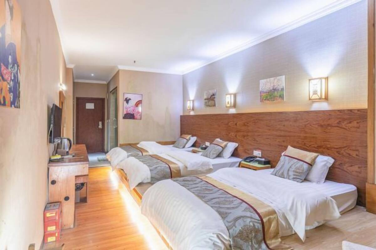 A hotel room with three beds, wooden headboard, wall-mounted lamps, and artwork.