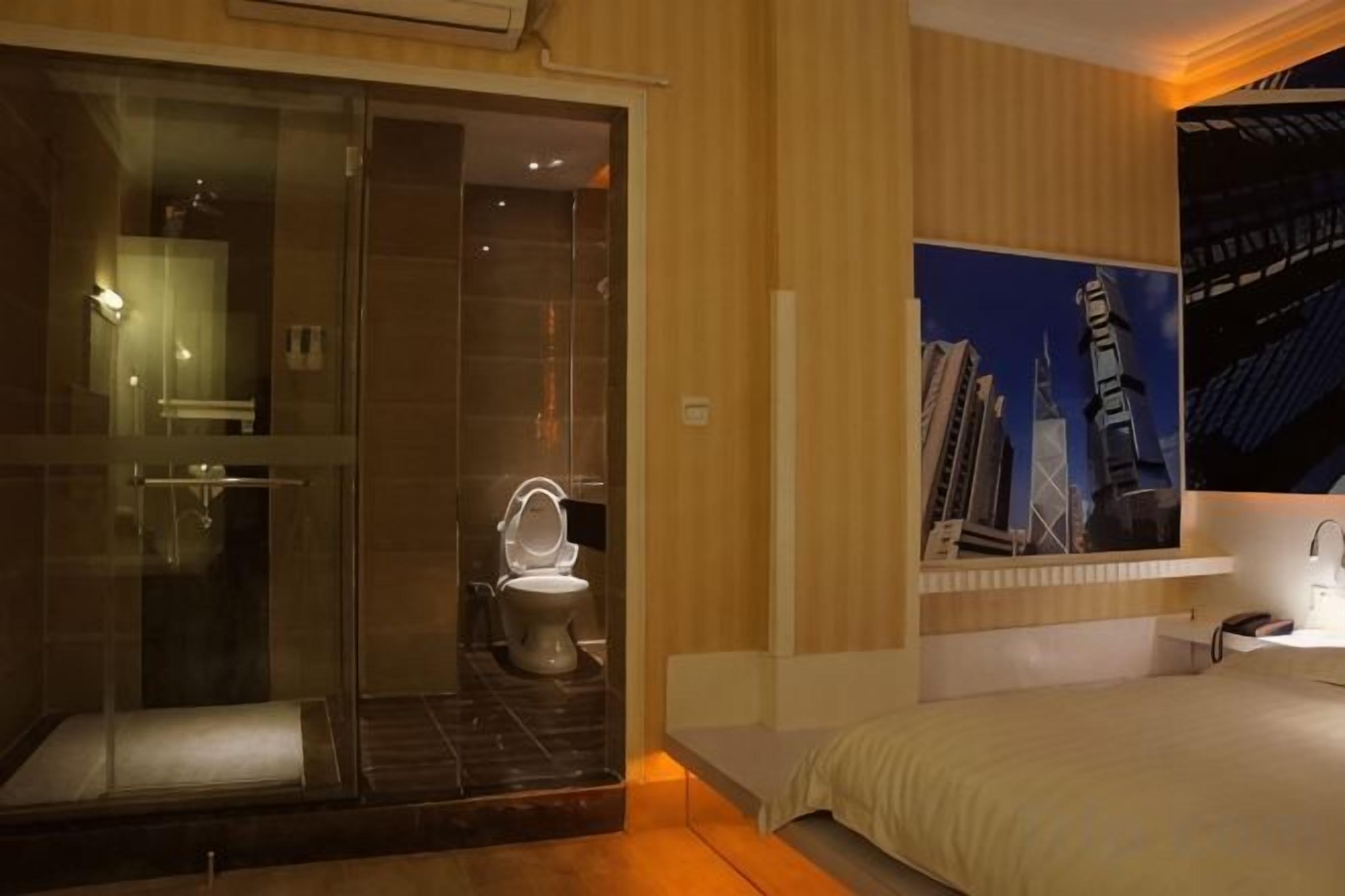 Habitación de hotel con ducha envuelta en vidrio, inodoro, cama y estante con pintura de un paisaje urbano.