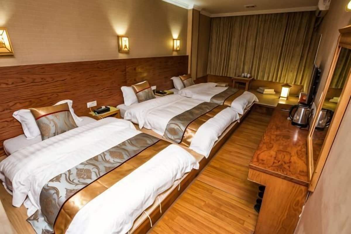 A hotel room with three beds, wooden flooring, a wooden headboard, a bedside table with a lamp, and a large mirror.