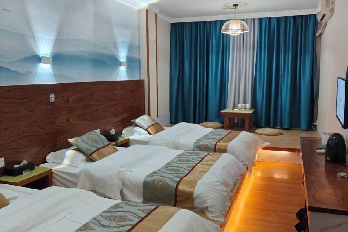 A hotel room with three beds, a wooden headboard, a desk with a computer, a television, and a view of the sky through a window.