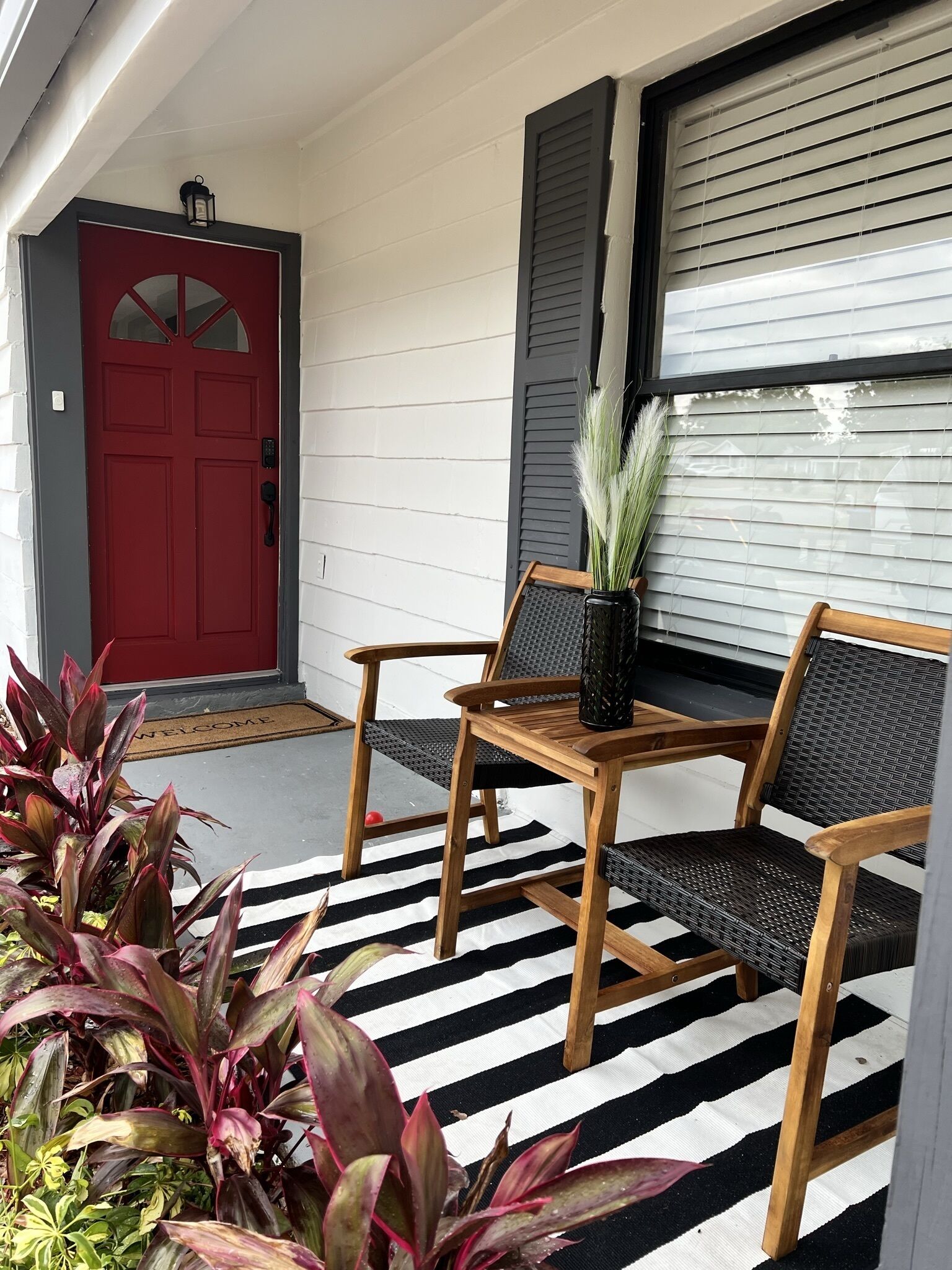 Un porche avec une porte rouge, deux chaises en bois, une petite table et une plante en pot.