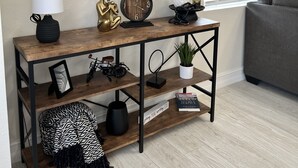 Un soggiorno con un tavolo consolle in legno, uno specchio rotondo, una lampada, una sveglia, oggetti decorativi e una finestra con persiane.