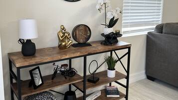 A living room with a wooden console table, a round mirror, a lamp, a clock, decorative items, and a window with blinds.