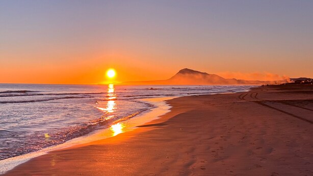 Una playa con un atardecer, olas y una montaña al fondo.