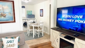 Una sala de estar con un televisor mostrando un juego de póker, un sofá con un cojín, un comedor con sillas y una cocina con lavadora.