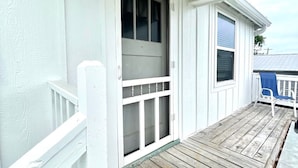Un porche blanco con una terraza de madera, una puerta blanca con un ventanal y una silla azul.