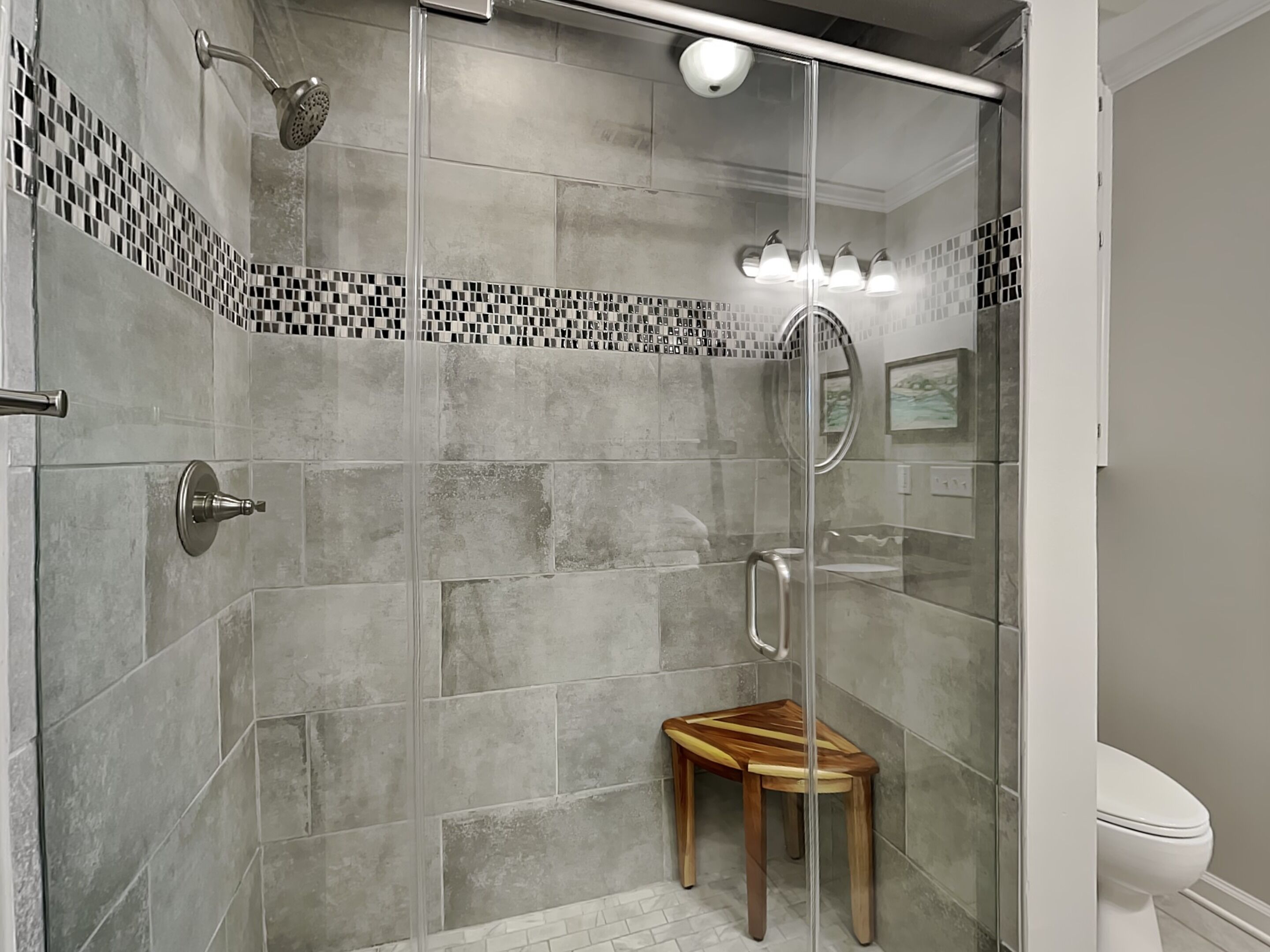 A modern shower with a glass door, a wooden stool, and a tiled floor.